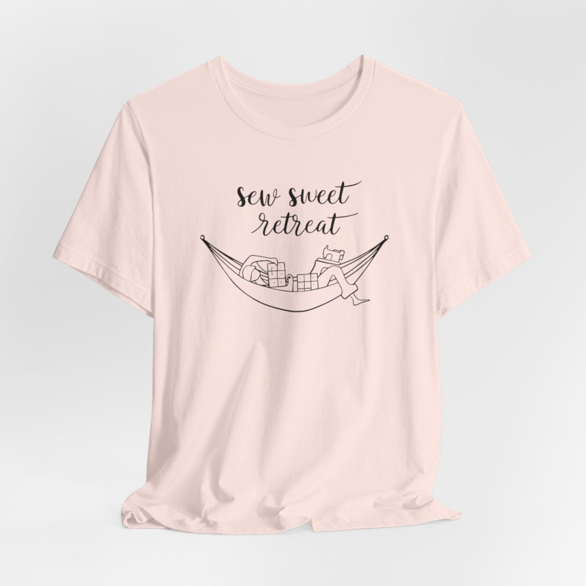 A Soft Pink T-Shirt with a black and white illustration of a hammock filled with quilting supplies like fabric squares and a sewing machine. Text above reads "Sew Sweet Retreat" in a flowing script font.