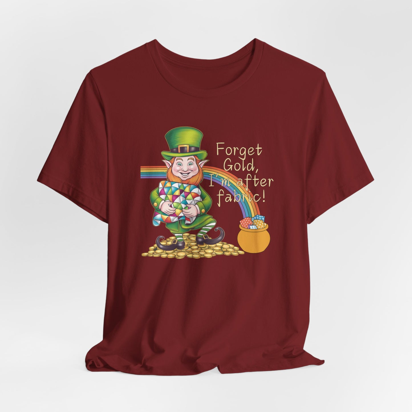 A Cardinal short-sleeve t-shirt featuring a cheerful leprechaun holding a quilt, sitting on gold coins, with a rainbow leading to a fabric-filled pot and the phrase 'Forget Gold, I’m After Fabric!' Perfect for quilters celebrating St. Patrick’s Day.