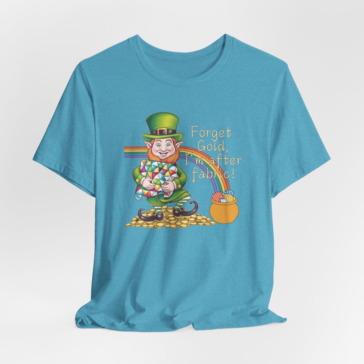 A Heather Aqua short-sleeve t-shirt featuring a cheerful leprechaun holding a quilt, sitting on gold coins, with a rainbow leading to a fabric-filled pot and the phrase 'Forget Gold, I’m After Fabric!' Perfect for quilters celebrating St. Patrick’s Day.