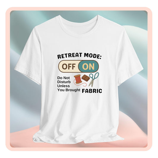 A A t-shirt with a toggle switch that says "Retreat Mode: OFF/ON" surrounded by a spool of thread, chocolate, and scissors. Text below reads "Do not disturb unless you brought fabric." Fun gift for quilting retreats.