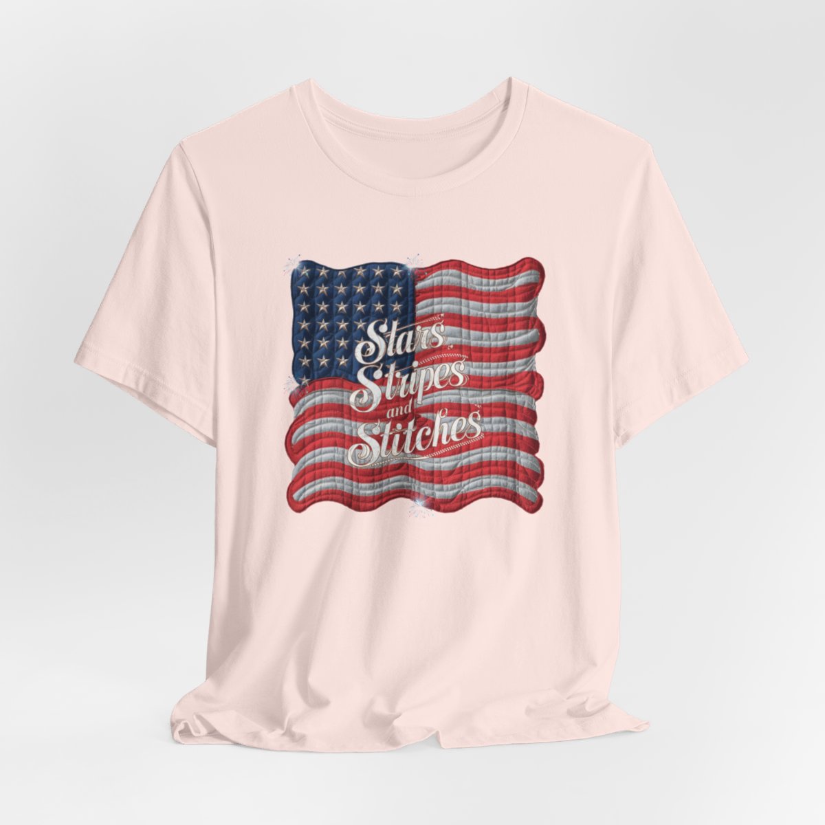 A Soft Pink T-shirt featuring quilted American flag design with text "Stars Stripes and Stitches" overlaid in stylized, patriotic font. Fireworks accent the corners.