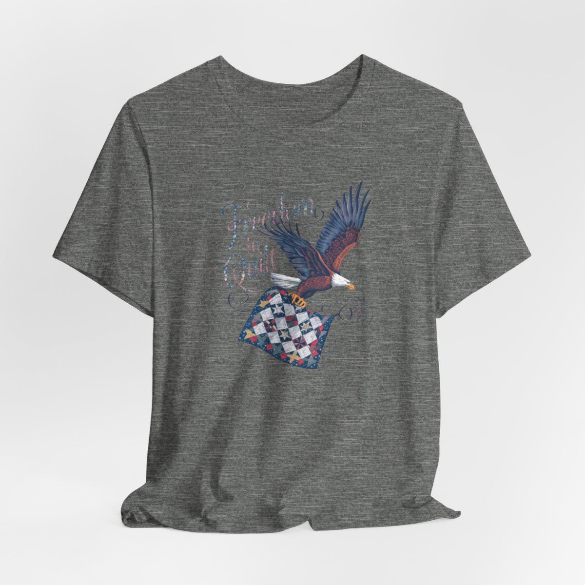 A Deep Heather T-shirt with detailed illustration of a bald eagle in flight, carrying a patchwork quilt. Text reads "Freedom to Quilt" in an ornate, patriotic font.