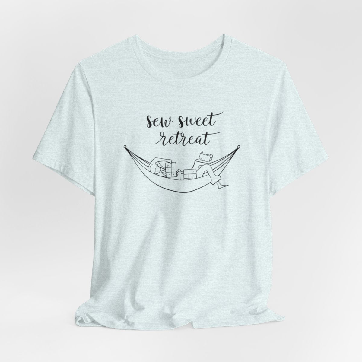 A Heather Ice Blue T-Shirt with a black and white illustration of a hammock filled with quilting supplies like fabric squares and a sewing machine. Text above reads "Sew Sweet Retreat" in a flowing script font.