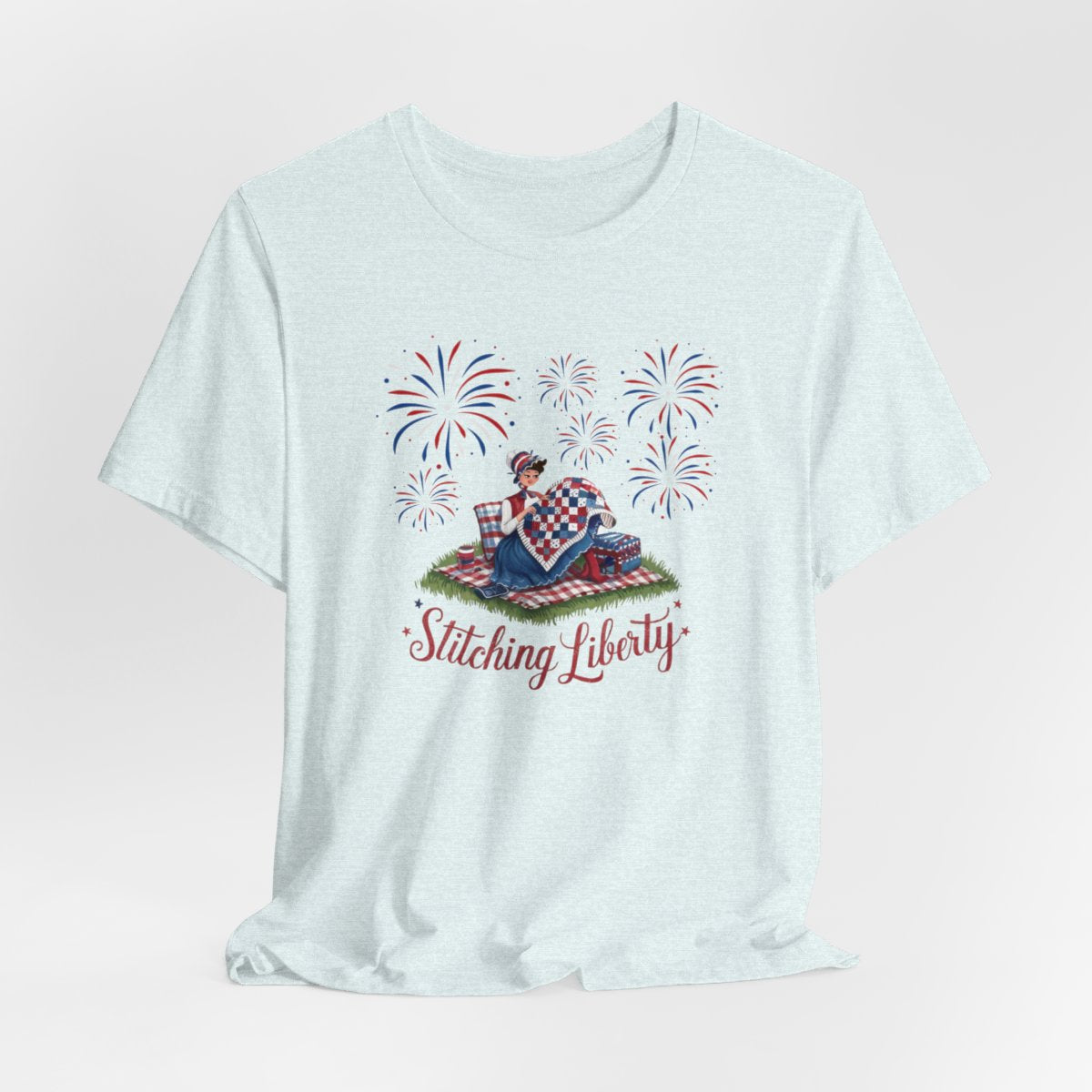 A Heather Ice Blue T-shirt with illustrated scene of a colonial woman quilting a patriotic blanket, surrounded by fireworks. Text reads "Stitching Liberty" in a decorative font.