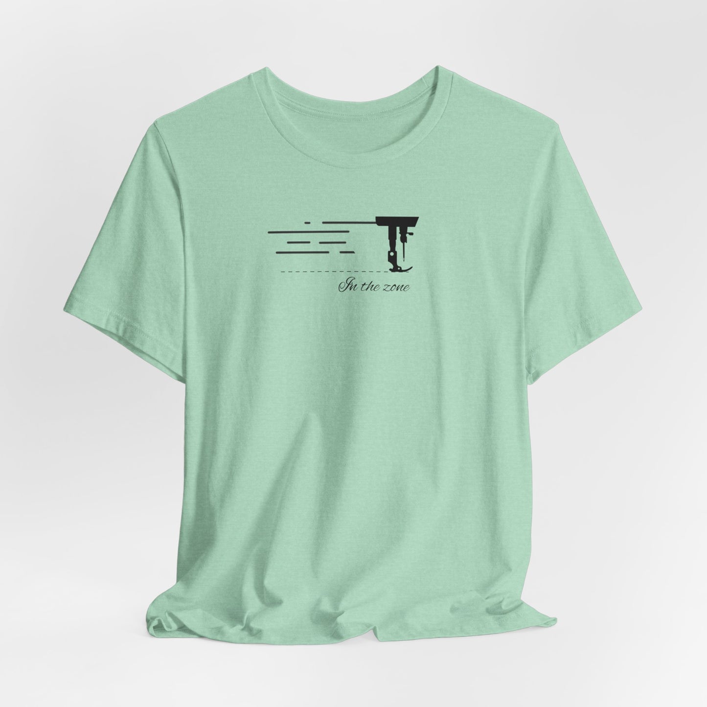 A Heather Mint T-shirt featuring a minimalist black silhouette of a sewing machine arm with motion lines. Below, the phrase "In the zone" is written in cursive script. The design conveys the focus and rhythm of quilting.