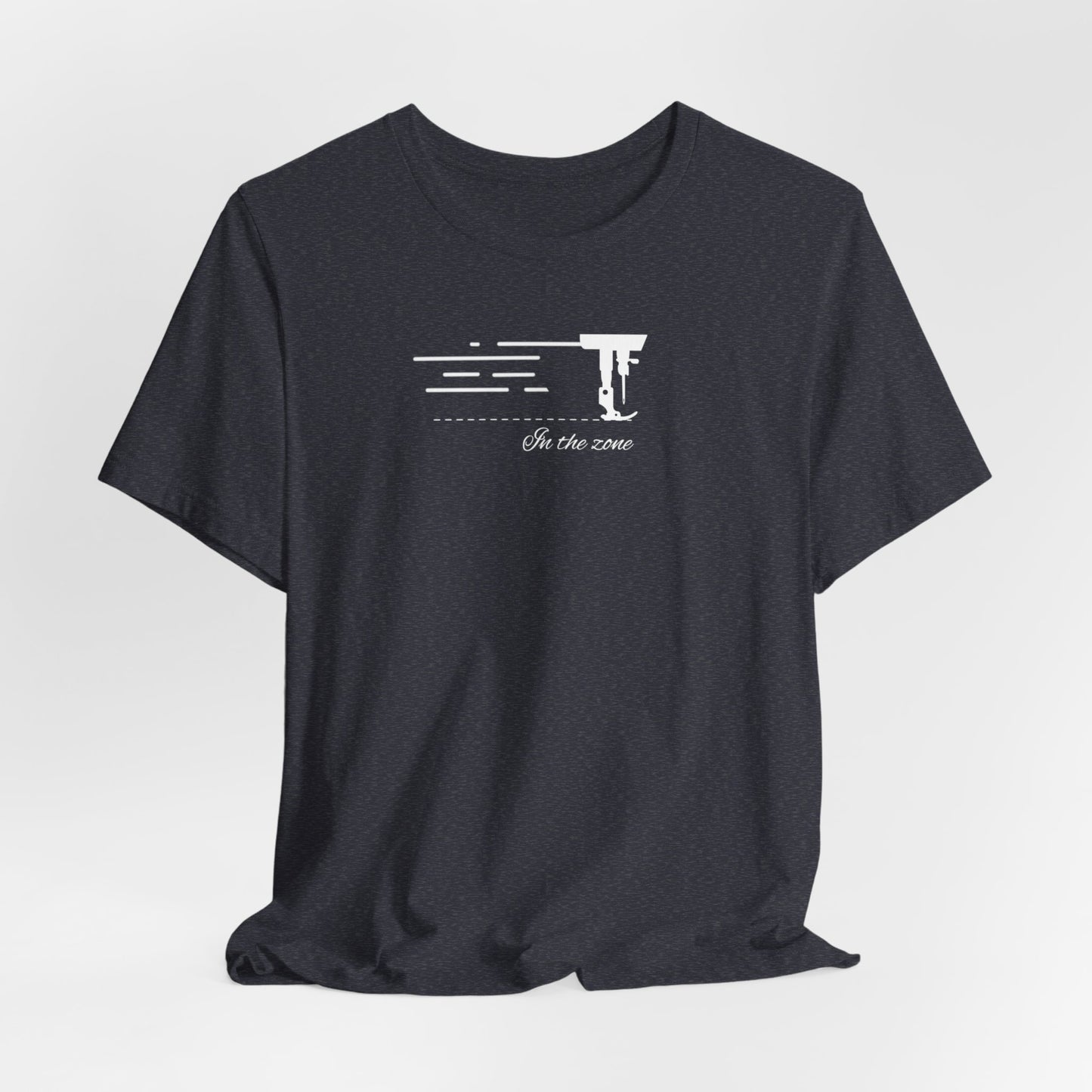 A Heather Navy T-shirt featuring a minimalist black silhouette of a sewing machine arm with motion lines. Below, the phrase "In the zone" is written in cursive script. The design conveys the focus and rhythm of quilting.