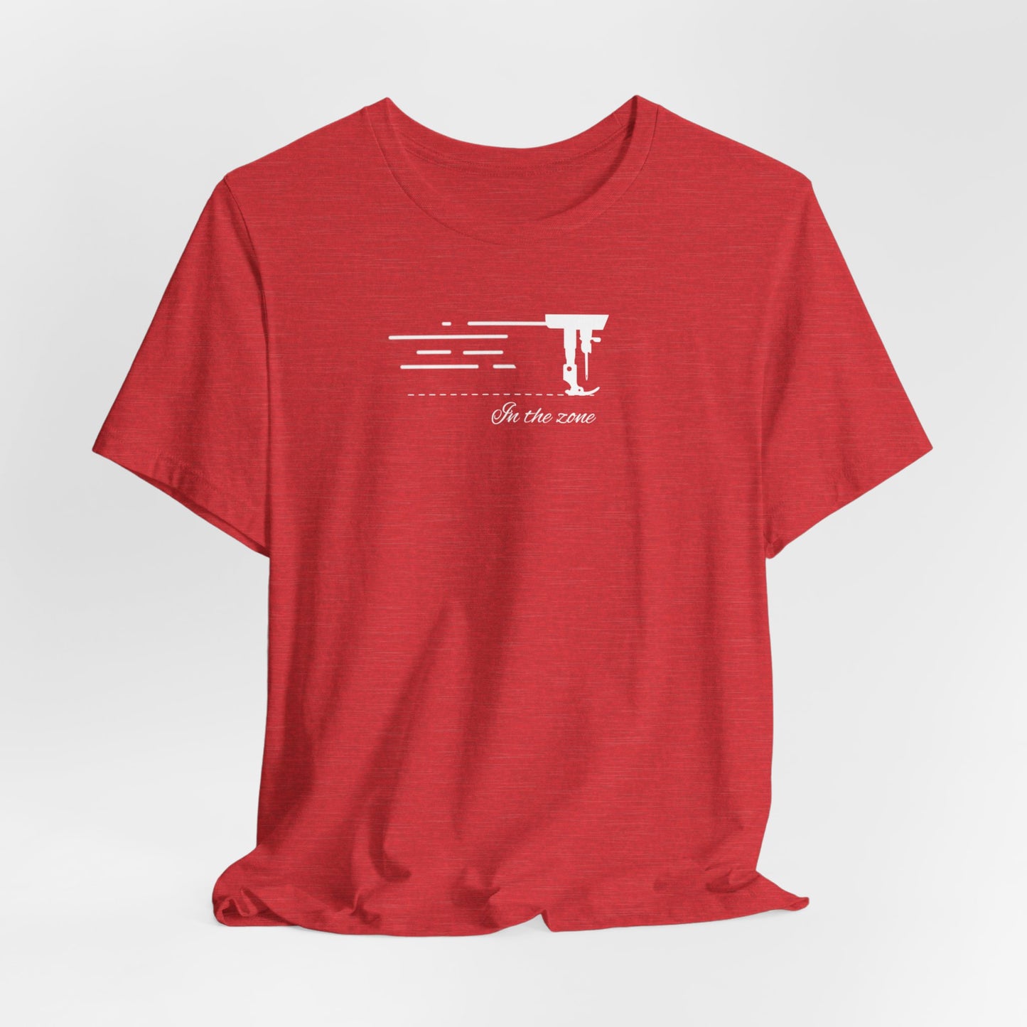 A Heather Red T-shirt featuring a minimalist black silhouette of a sewing machine arm with motion lines. Below, the phrase "In the zone" is written in cursive script. The design conveys the focus and rhythm of quilting.