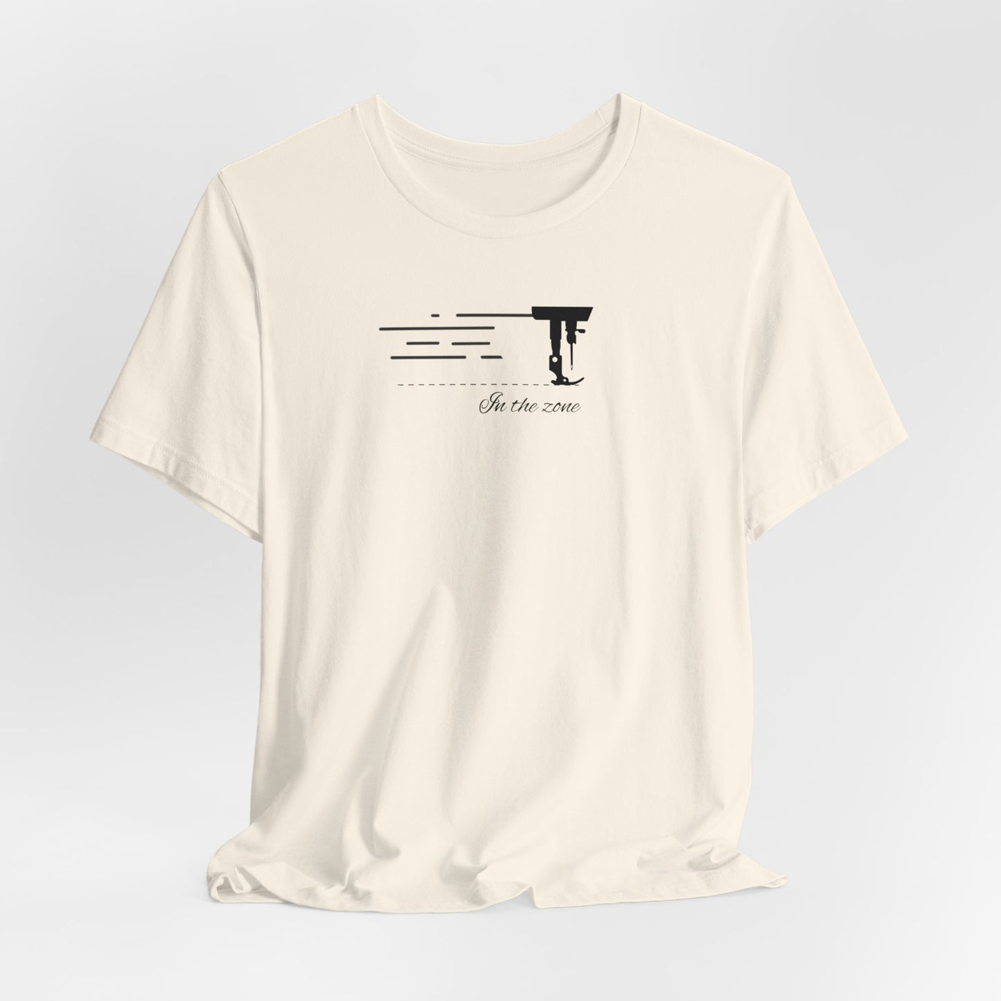 A Natural T-shirt featuring a minimalist black silhouette of a sewing machine arm with motion lines. Below, the phrase "In the zone" is written in cursive script. The design conveys the focus and rhythm of quilting.