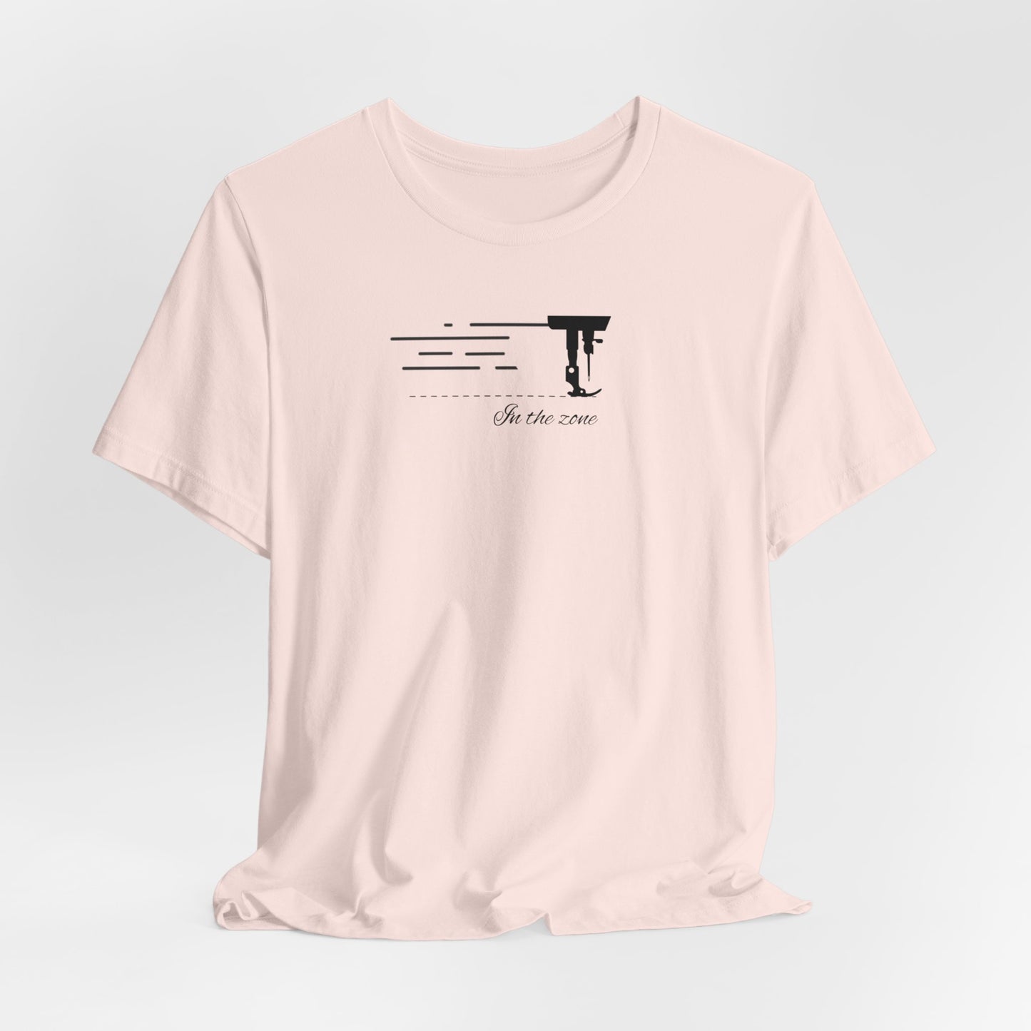 A Soft Pink T-shirt featuring a minimalist black silhouette of a sewing machine arm with motion lines. Below, the phrase "In the zone" is written in cursive script. The design conveys the focus and rhythm of quilting.