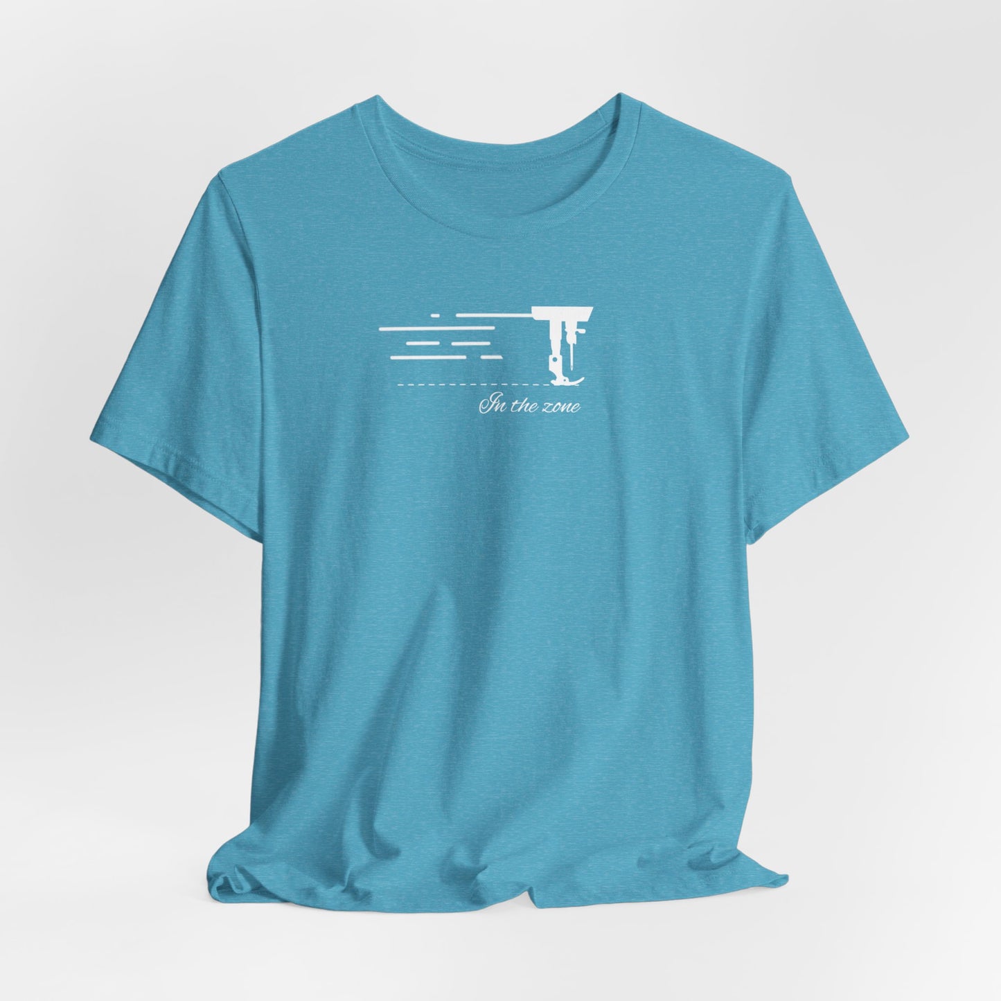 A Heather Aqua T-shirt featuring a minimalist black silhouette of a sewing machine arm with motion lines. Below, the phrase "In the zone" is written in cursive script. The design conveys the focus and rhythm of quilting.