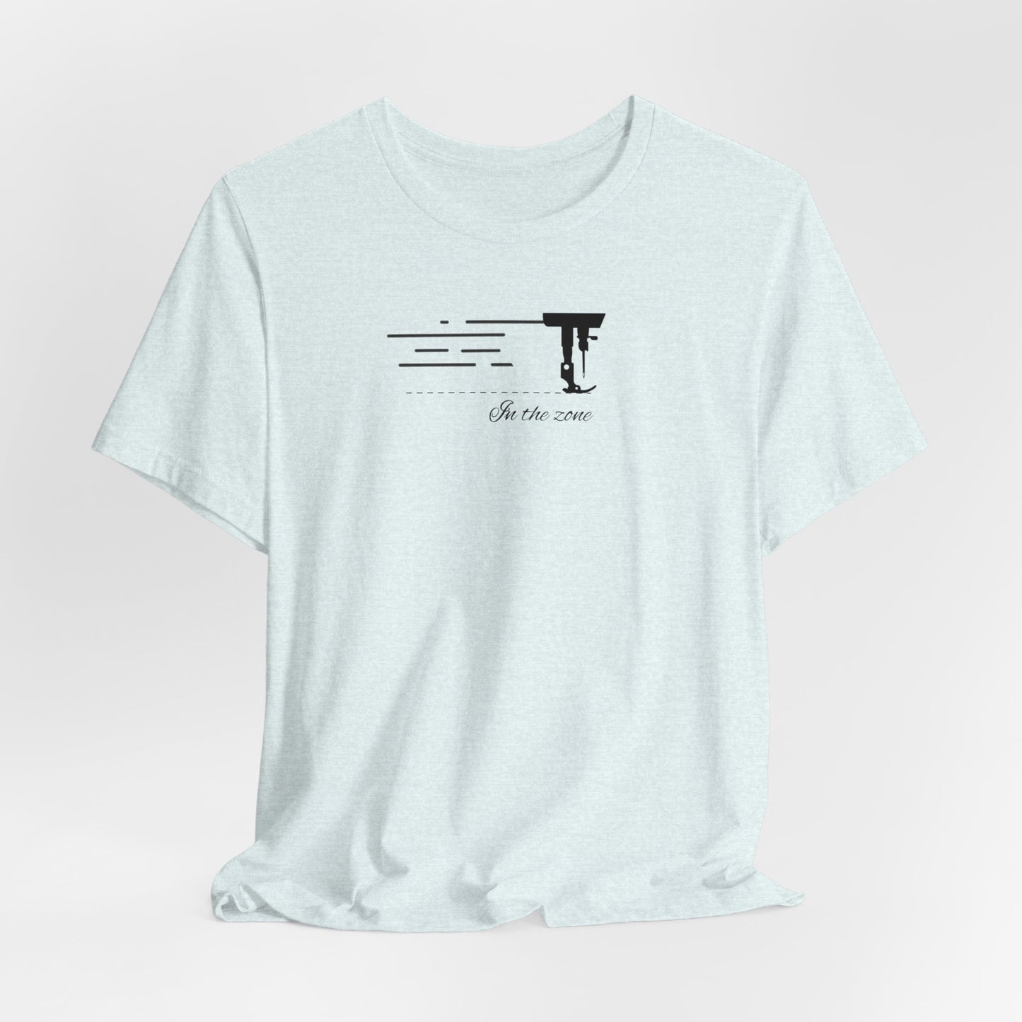 A Heather Ice Blue T-shirt featuring a minimalist black silhouette of a sewing machine arm with motion lines. Below, the phrase "In the zone" is written in cursive script. The design conveys the focus and rhythm of quilting.