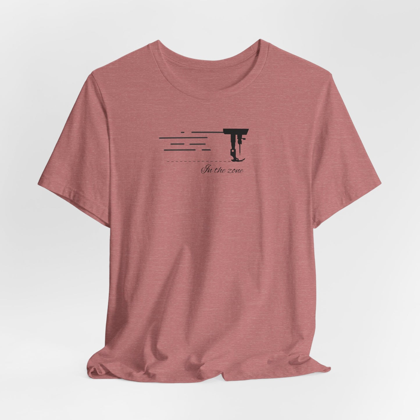 A Heather Mauve T-shirt featuring a minimalist black silhouette of a sewing machine arm with motion lines. Below, the phrase "In the zone" is written in cursive script. The design conveys the focus and rhythm of quilting.