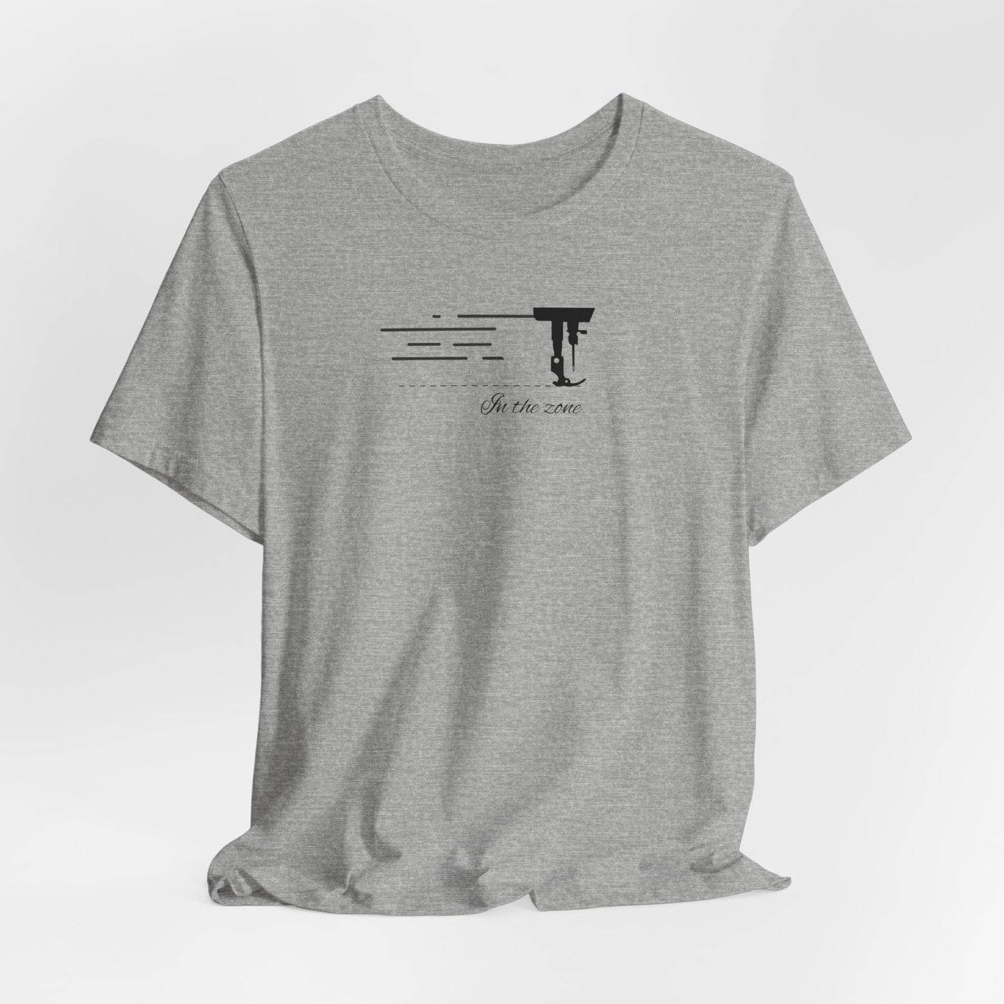 An Athletic Heather T-shirt featuring a minimalist black silhouette of a sewing machine arm with motion lines. Below, the phrase "In the zone" is written in cursive script. The design conveys the focus and rhythm of quilting.