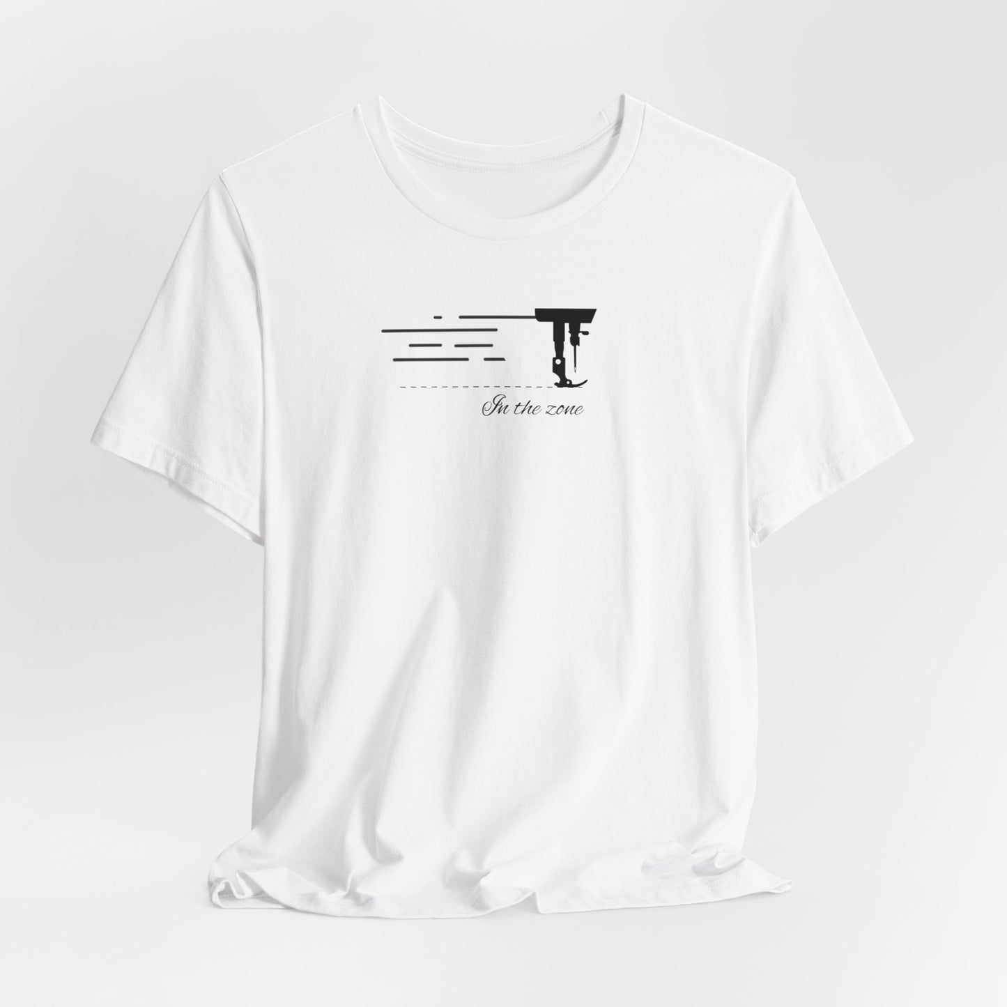 A White T-shirt featuring a minimalist black silhouette of a sewing machine arm with motion lines. Below, the phrase "In the zone" is written in cursive script. The design conveys the focus and rhythm of quilting.