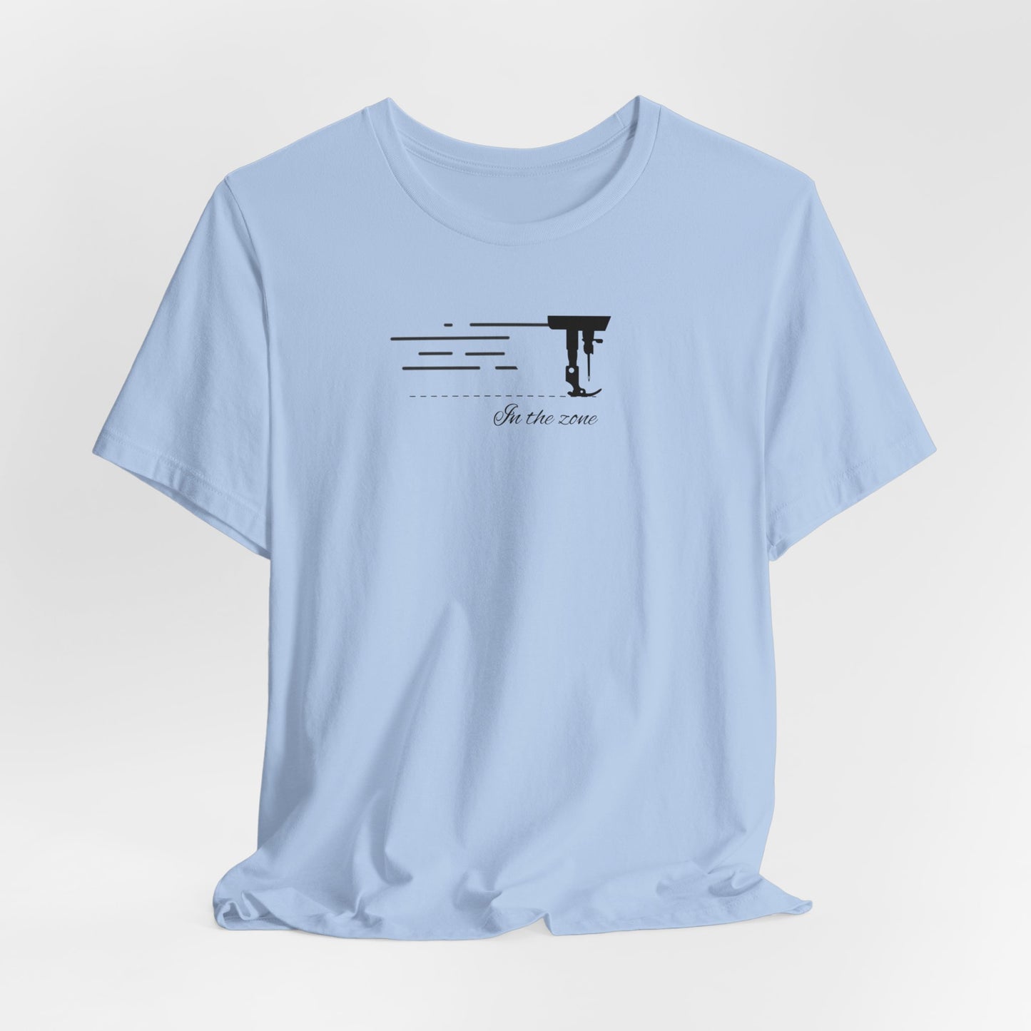 A Baby Blue T-shirt featuring a minimalist black silhouette of a sewing machine arm with motion lines. Below, the phrase "In the zone" is written in cursive script. The design conveys the focus and rhythm of quilting.