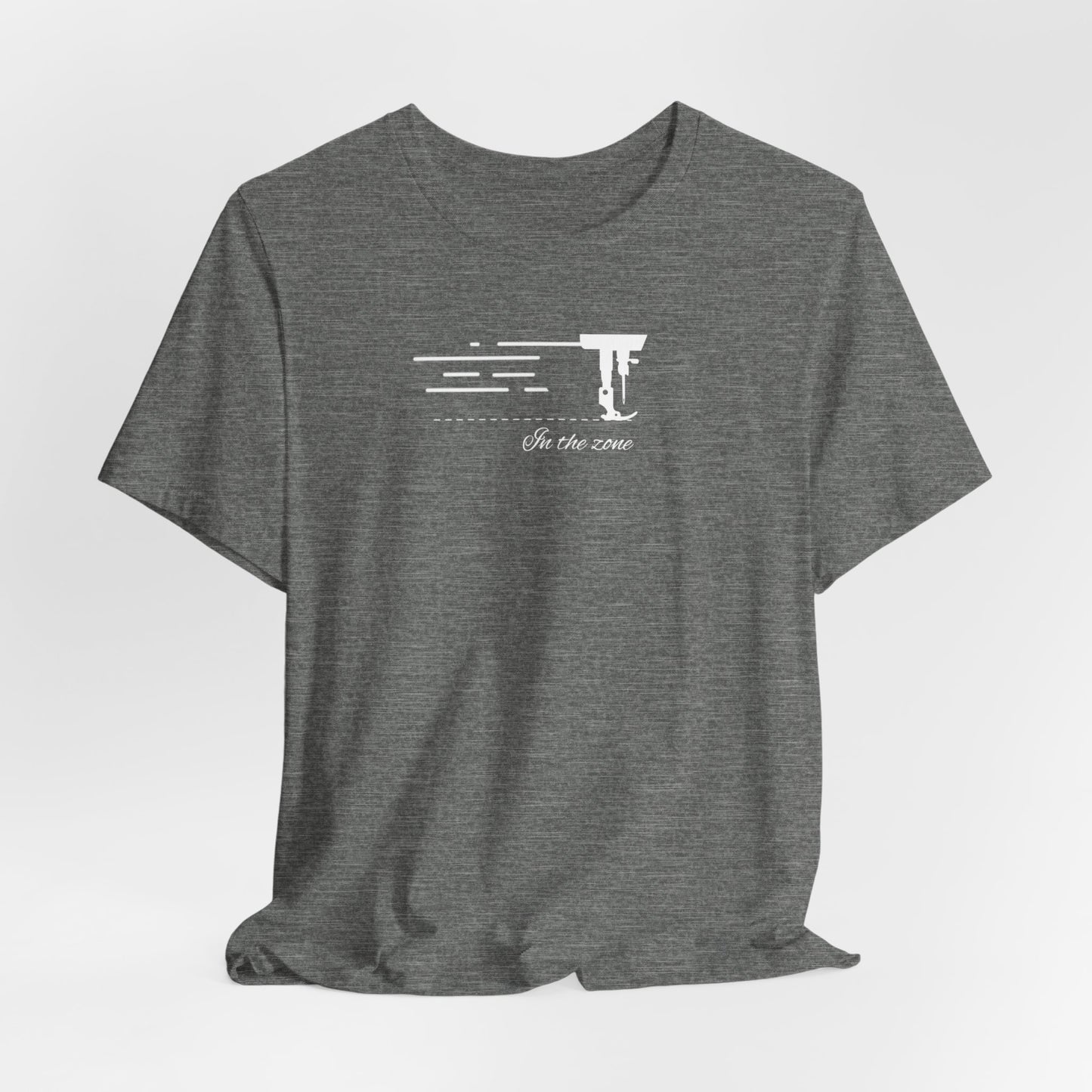 A Deep Heather T-shirt featuring a minimalist black silhouette of a sewing machine arm with motion lines. Below, the phrase "In the zone" is written in cursive script. The design conveys the focus and rhythm of quilting.