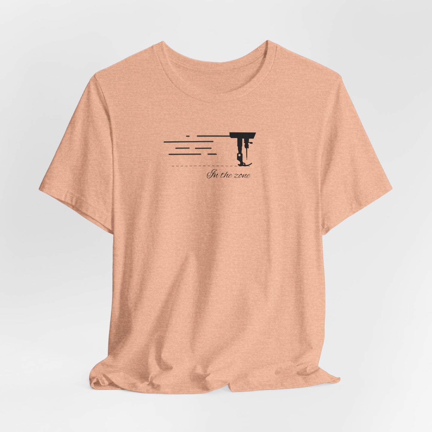 A Heather Peach T-shirt featuring a minimalist black silhouette of a sewing machine arm with motion lines. Below, the phrase "In the zone" is written in cursive script. The design conveys the focus and rhythm of quilting.