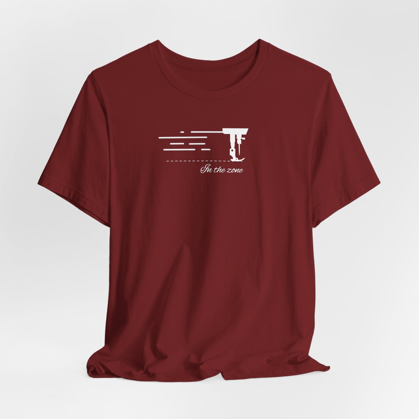 A Cardinal T-shirt featuring a minimalist black silhouette of a sewing machine arm with motion lines. Below, the phrase "In the zone" is written in cursive script. The design conveys the focus and rhythm of quilting.