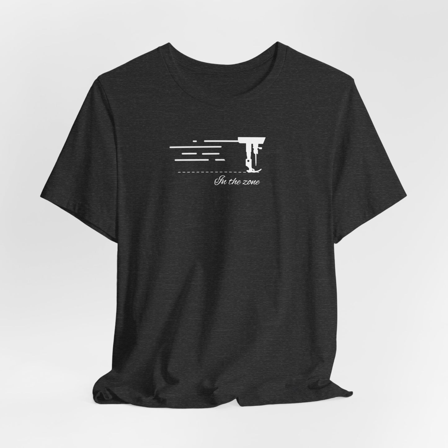 A Dark Grey Heather T-shirt featuring a minimalist black silhouette of a sewing machine arm with motion lines. Below, the phrase "In the zone" is written in cursive script. The design conveys the focus and rhythm of quilting.