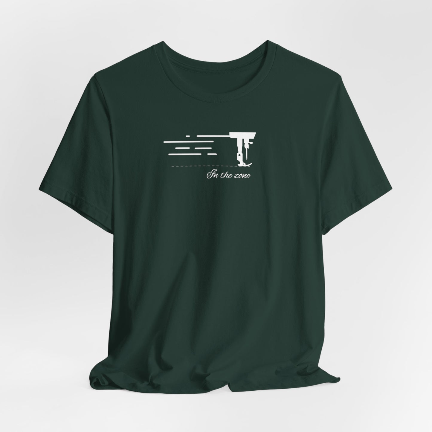 A Forest T-shirt featuring a minimalist black silhouette of a sewing machine arm with motion lines. Below, the phrase "In the zone" is written in cursive script. The design conveys the focus and rhythm of quilting.