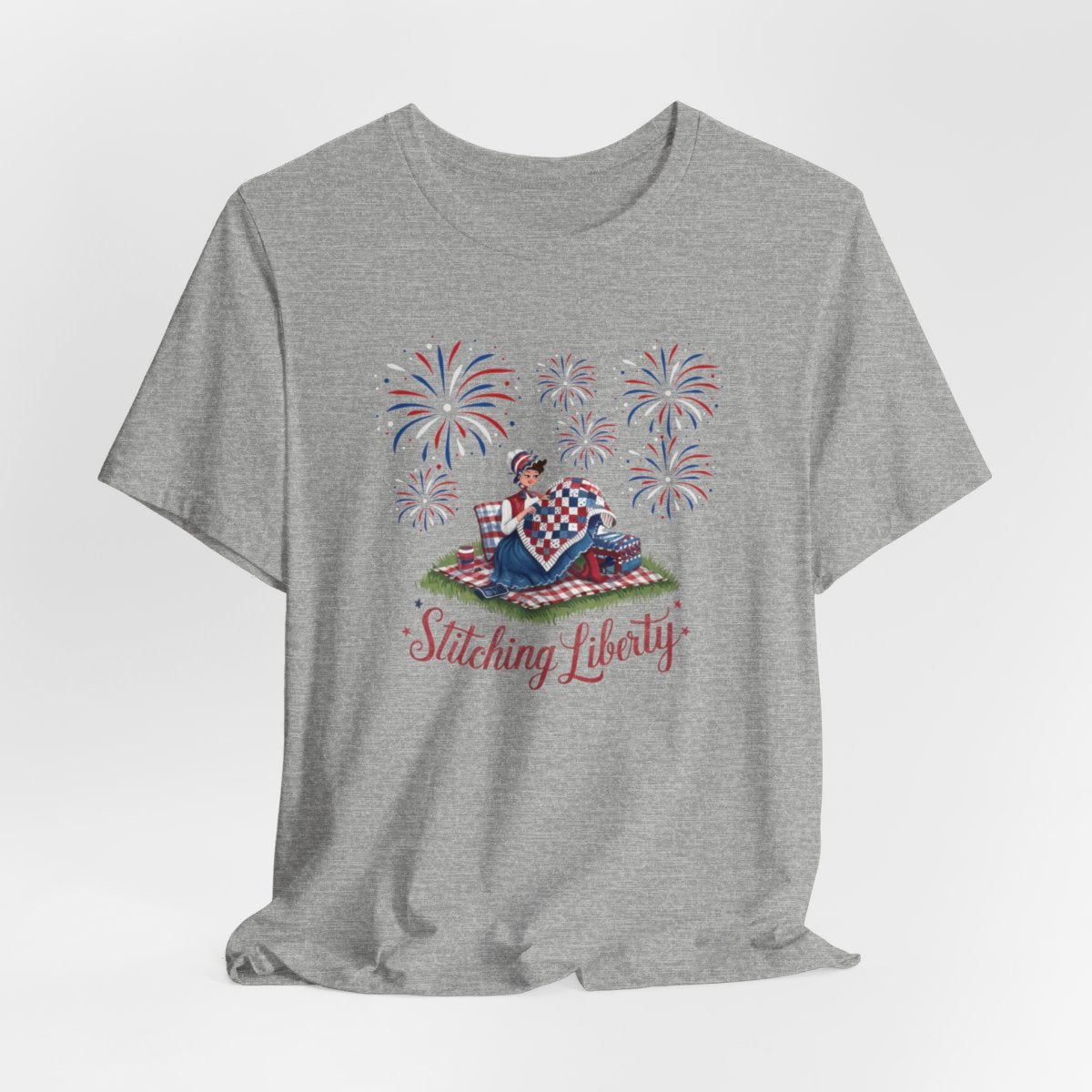 An Athletic Heather T-shirt with illustrated scene of a colonial woman quilting a patriotic blanket, surrounded by fireworks. Text reads "Stitching Liberty" in a decorative font.