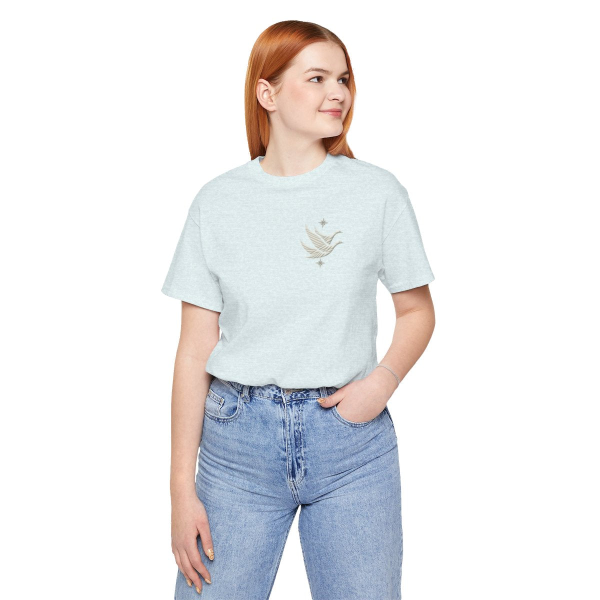 A Heather Ice Blue T-shirt featuring two stylized white geese in flight, with outstretched wings. The geese are composed of intricate geometric patterns resembling quilt stitching. Two eight-pointed stars frame the scene, one above and one below the geese, adding a celestial element to the peaceful composition.