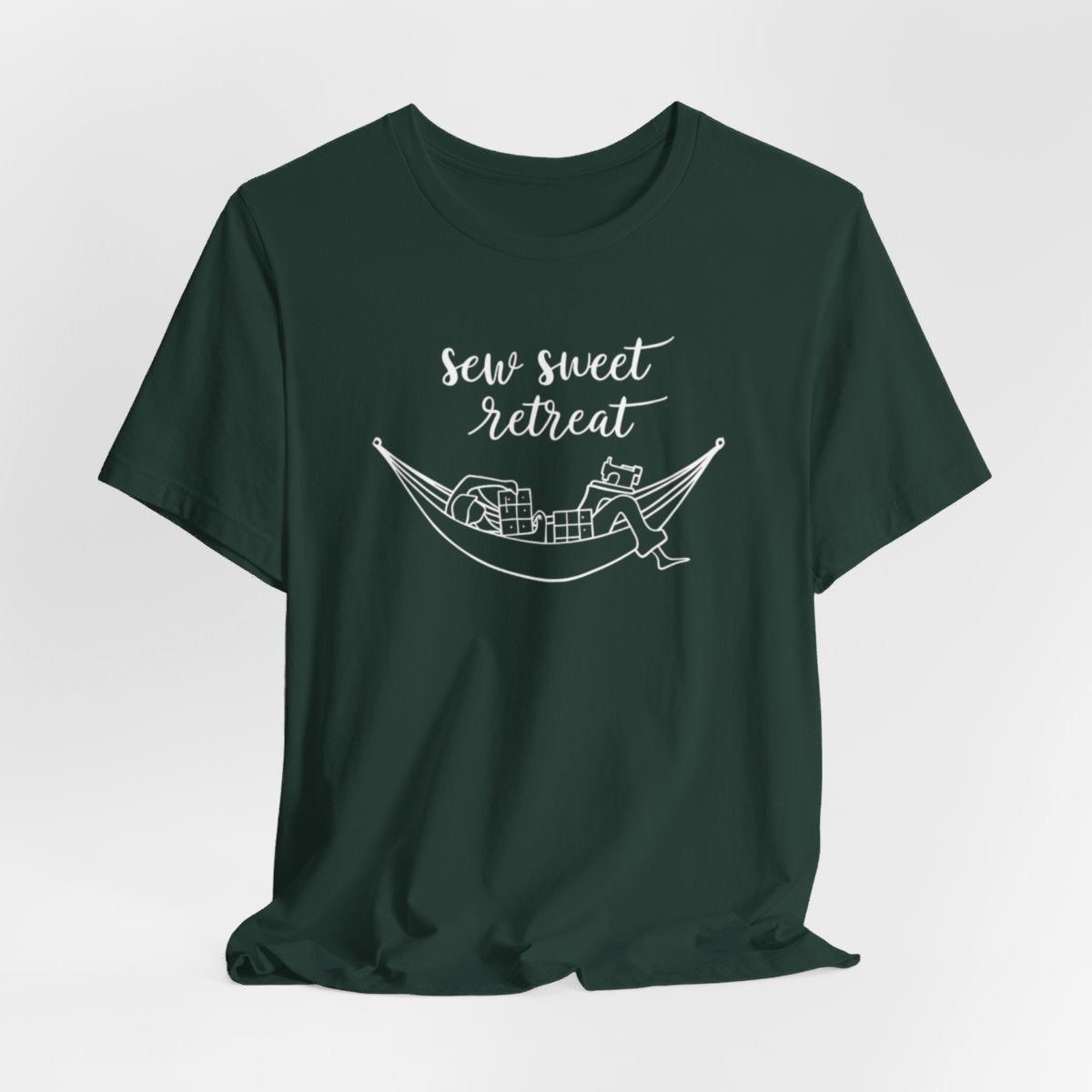 A Forest T-Shirt with a black and white illustration of a hammock filled with quilting supplies like fabric squares and a sewing machine. Text above reads "Sew Sweet Retreat" in a flowing script font.