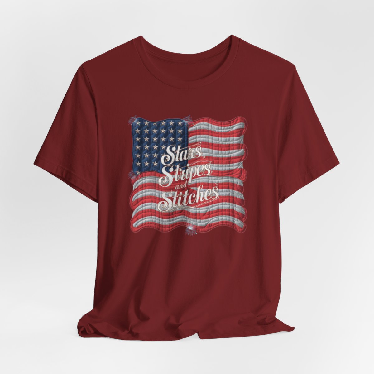 A Cardinal T-shirt featuring quilted American flag design with text "Stars Stripes and Stitches" overlaid in stylized, patriotic font. Fireworks accent the corners.