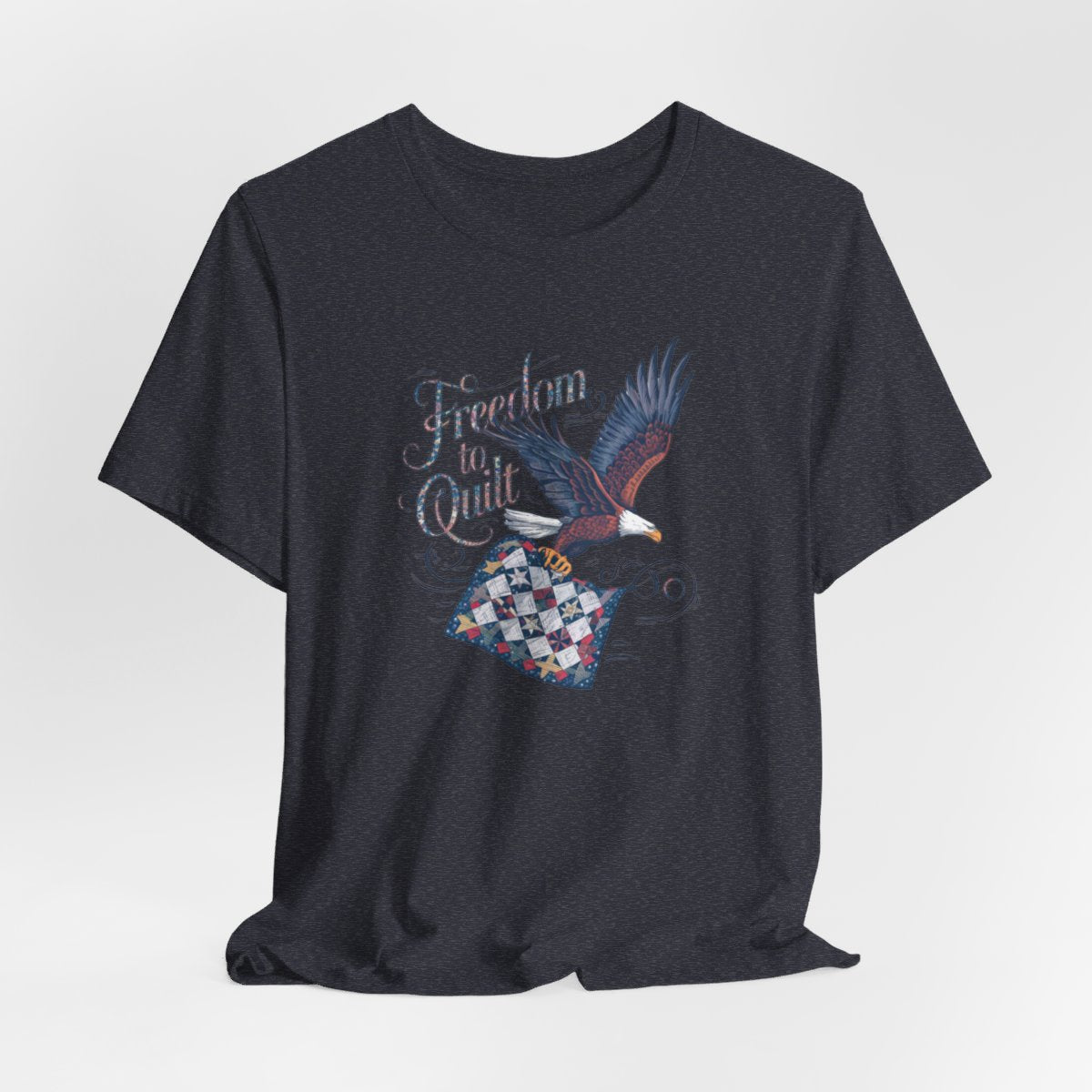 A Heather Navy T-shirt with detailed illustration of a bald eagle in flight, carrying a patchwork quilt. Text reads "Freedom to Quilt" in an ornate, patriotic font.