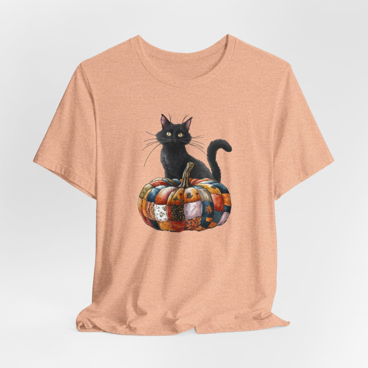 A Heather Peach Halloween Quilting T-Shirt featuring a black cat sitting on colorful patchwork quilt pumpkin. Cat has yellow eyes and long whiskers. Pumpkin features various quilt patterns in autumn colors. Halloween-themed t-shirt design for quilting enthusiasts.