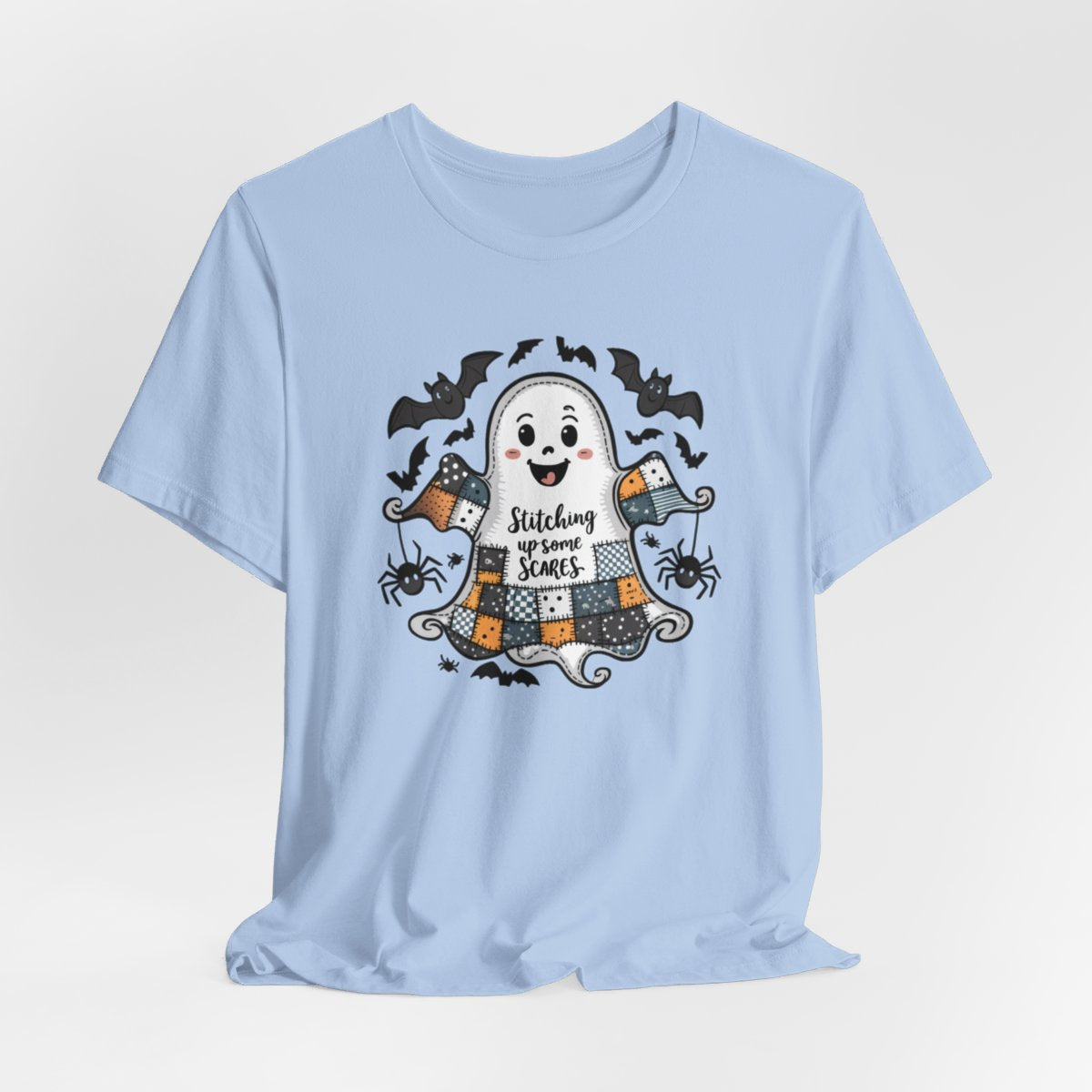 A Baby Blue Halloween Quilting T-Shirt featuring a smiling ghost with quilted patchwork body. Halloween-themed quilt patterns. Cute bats and spiders around ghost. Text reads "Stitching up some Scares". Ideal for quilters who enjoy Halloween themes.