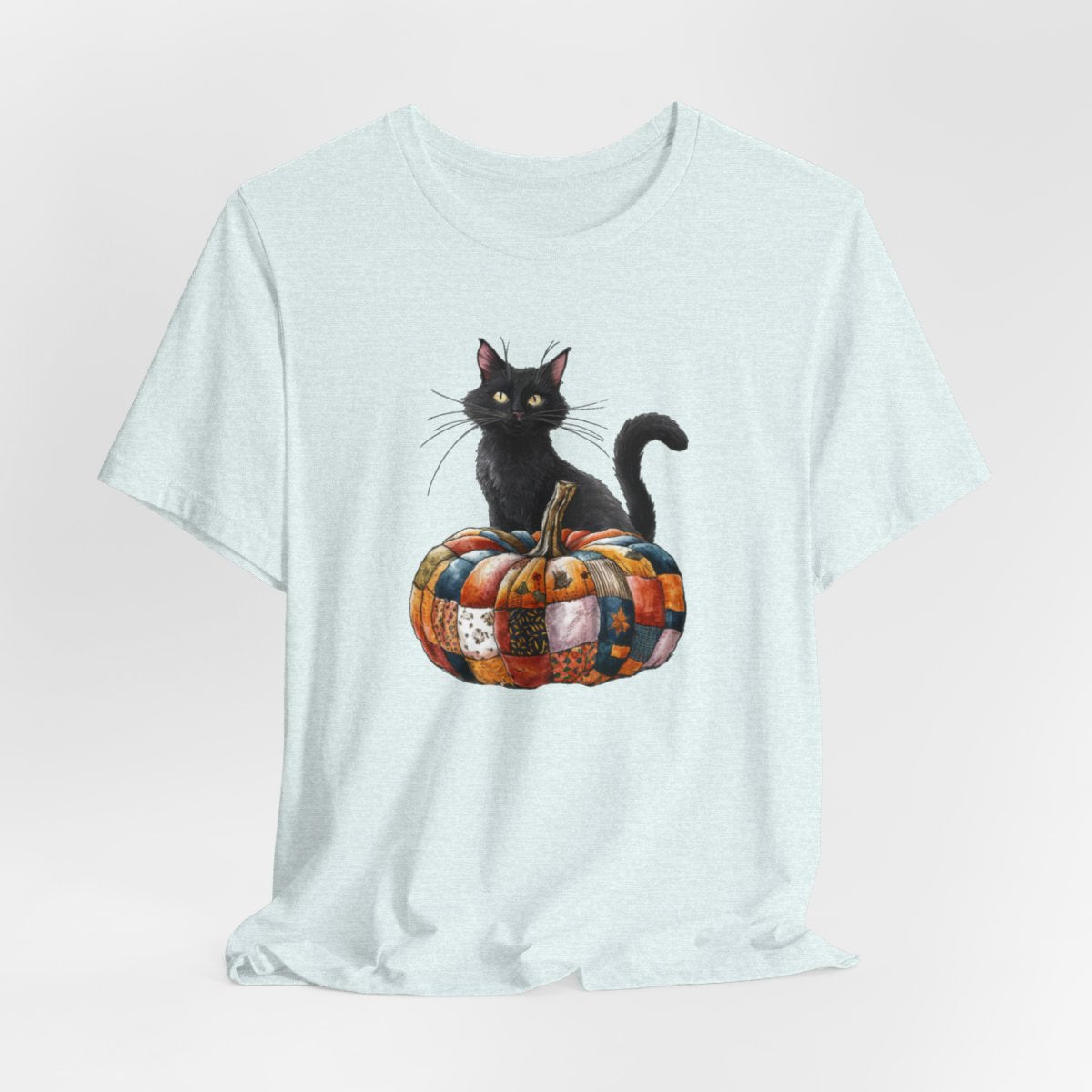 A Heather Ice Blue Halloween Quilting T-Shirt featuring a black cat sitting on colorful patchwork quilt pumpkin. Cat has yellow eyes and long whiskers. Pumpkin features various quilt patterns in autumn colors. Halloween-themed t-shirt design for quilting enthusiasts.