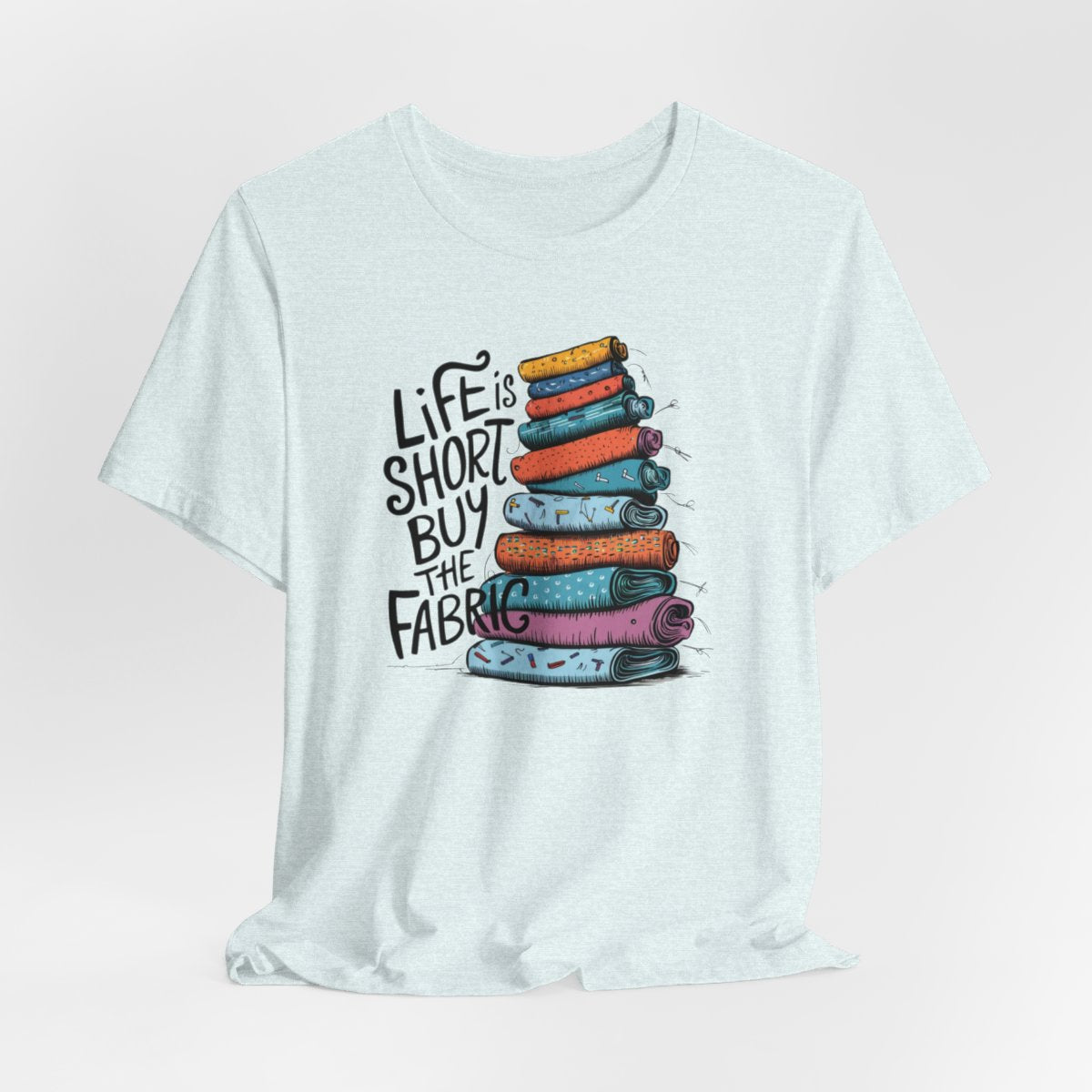 A Heather Ice Blue fun T-Shirt with the phrase Life is Short, Buy the Fabric with the label 'Life is Short, Buy the Fabric' next to a graphic of a fun pile of fabric