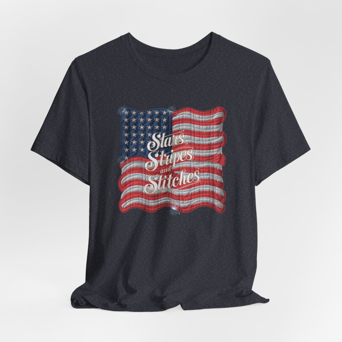 A Heather Navy T-shirt featuring quilted American flag design with text "Stars Stripes and Stitches" overlaid in stylized, patriotic font. Fireworks accent the corners.