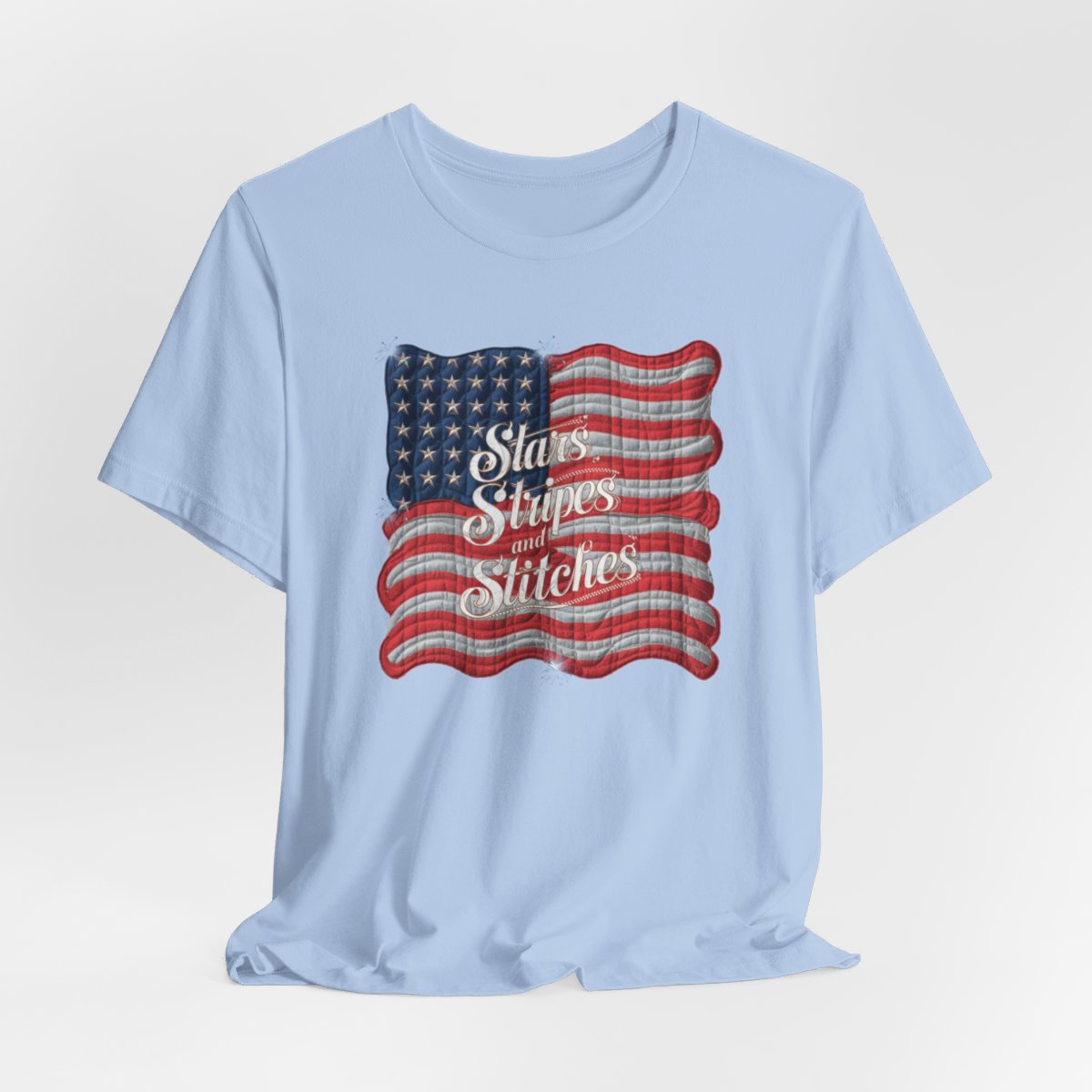 A Baby Blue T-shirt featuring quilted American flag design with text "Stars Stripes and Stitches" overlaid in stylized, patriotic font. Fireworks accent the corners.