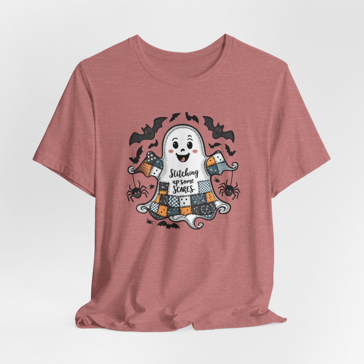 A Heather Mauve Halloween Quilting T-Shirt featuring a smiling ghost with quilted patchwork body. Halloween-themed quilt patterns. Cute bats and spiders around ghost. Text reads "Stitching up some Scares". Ideal for quilters who enjoy Halloween themes.