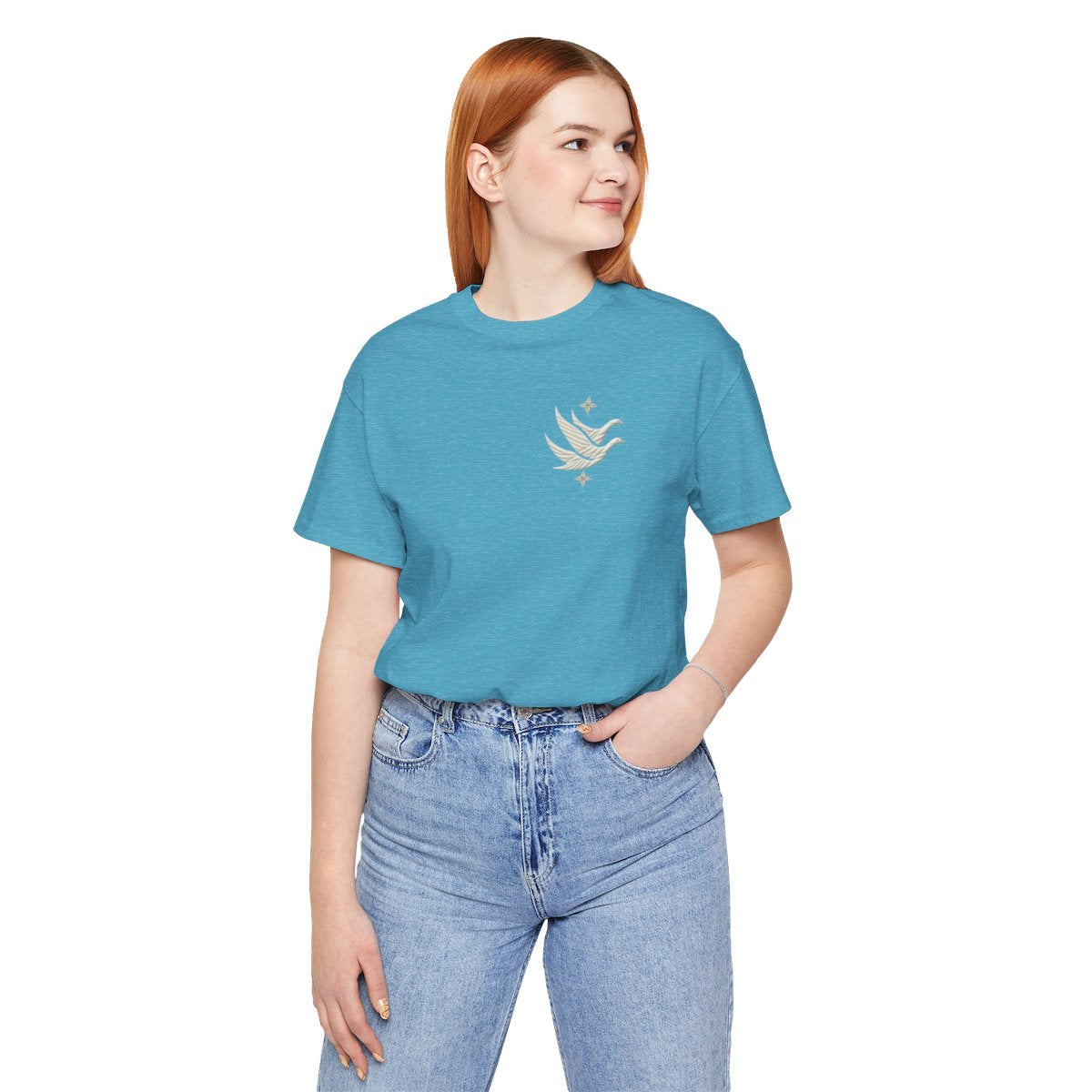 A Heather Aqua T-shirt featuring two stylized white geese in flight, with outstretched wings. The geese are composed of intricate geometric patterns resembling quilt stitching. Two eight-pointed stars frame the scene, one above and one below the geese, adding a celestial element to the peaceful composition.