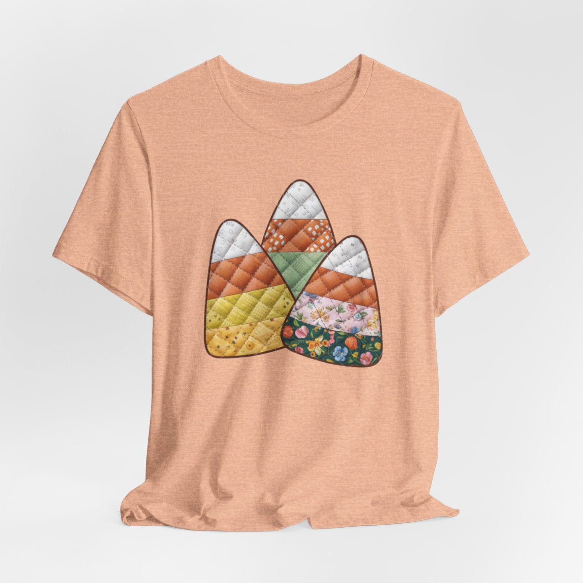 A Heather Peach Halloween Quilting T-Shirt featuring three large candy corn shapes made of quilted fabrics with various patterns. Each candy corn section features different quilting motifs in autumn colors. Playful take on Halloween candy and quilting craft.