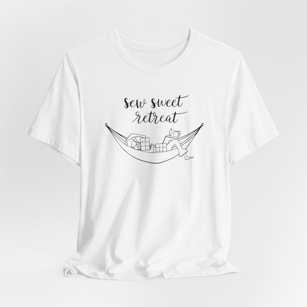 A White T-Shirt with a black and white illustration of a hammock filled with quilting supplies like fabric squares and a sewing machine. Text above reads "Sew Sweet Retreat" in a flowing script font.