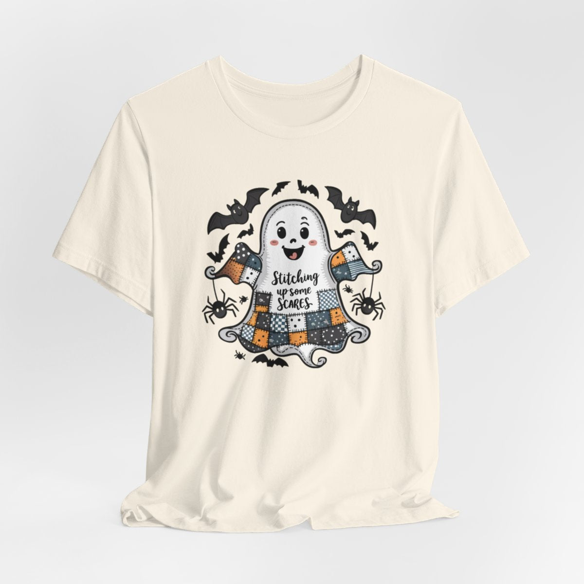 A Natural Halloween Quilting T-Shirt featuring a smiling ghost with quilted patchwork body. Halloween-themed quilt patterns. Cute bats and spiders around ghost. Text reads "Stitching up some Scares". Ideal for quilters who enjoy Halloween themes.