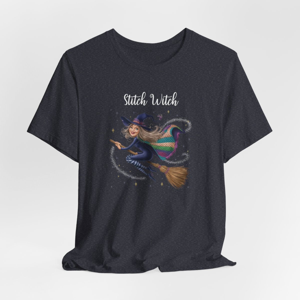 A Heather Navy Halloween Quilting T-Shirt featuring a witch riding broomstick on t-shirt design. Colorful patchwork quilt cape with various patterns. Sparkling magic and stars. Text reads "Stitch Witch". Ideal for quilters who enjoy magical themes.