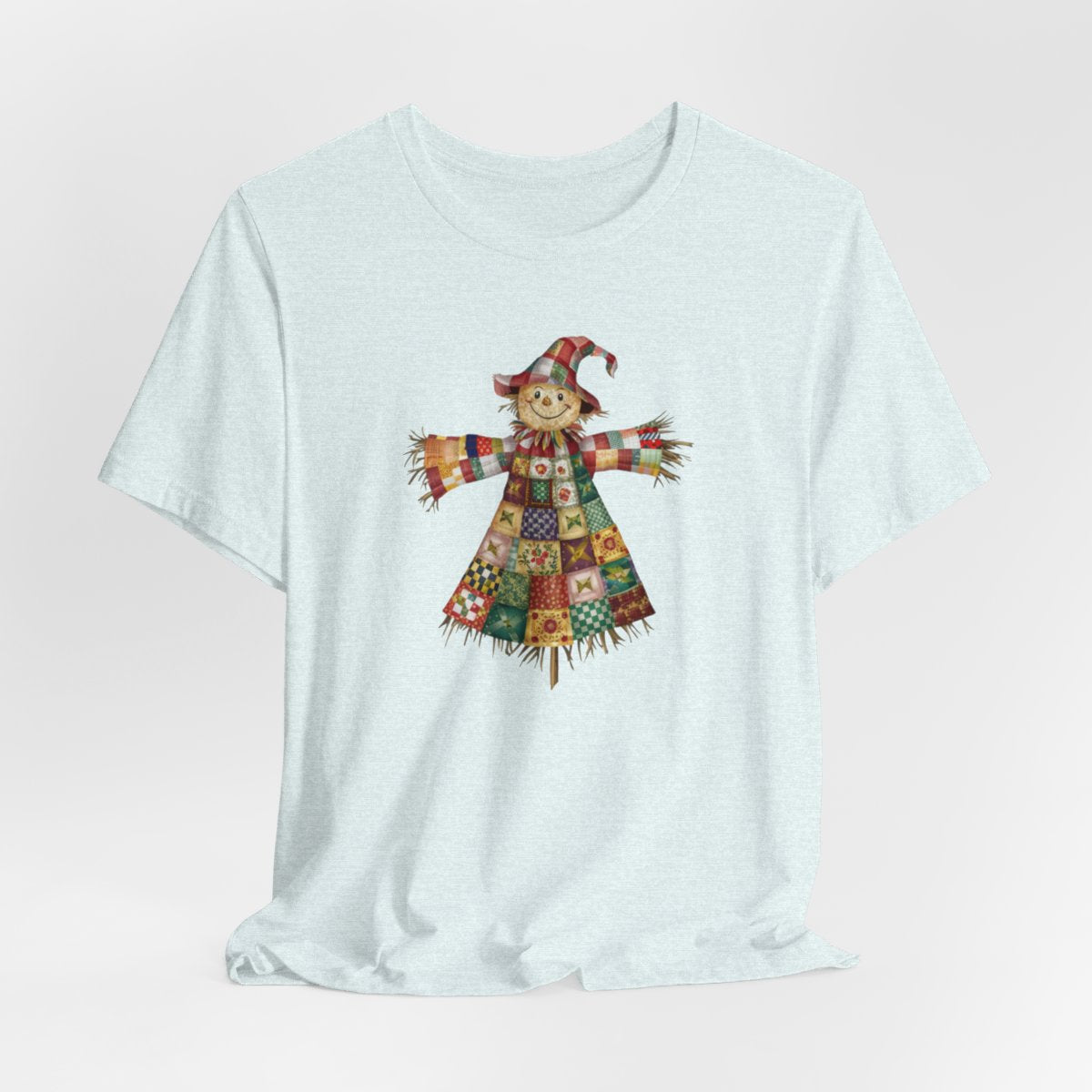 A Heather Ice Blue thanksgiving  quilting t-shirt featuring a smiling scarecrow made entirely of colorful quilt patterns. Wearing a patchwork hat and clothes with various quilt designs. Straw details on hands and neck. Cheerful autumn-themed illustration celebrating quilting craft.