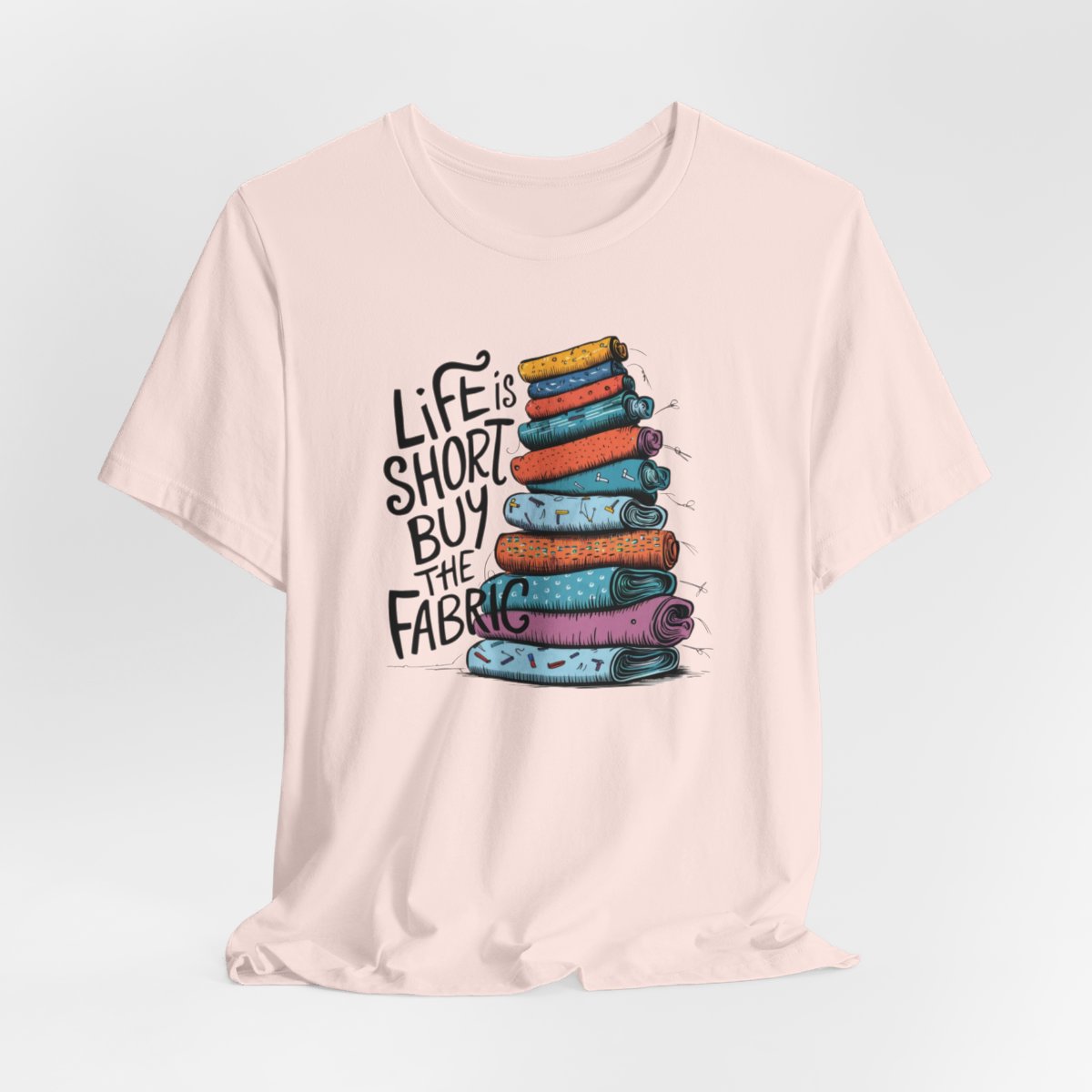 A Soft Pink fun T-Shirt with the phrase Life is Short, Buy the Fabric with the label 'Life is Short, Buy the Fabric' next to a graphic of a fun pile of fabric