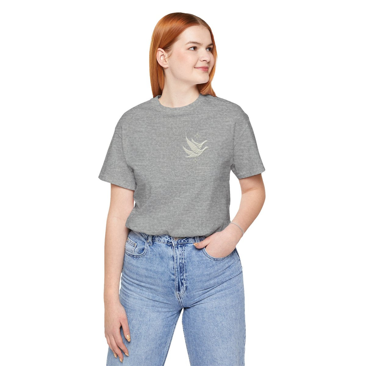 An Athletic Heather T-shirt featuring two stylized white geese in flight, with outstretched wings. The geese are composed of intricate geometric patterns resembling quilt stitching. Two eight-pointed stars frame the scene, one above and one below the geese, adding a celestial element to the peaceful composition.