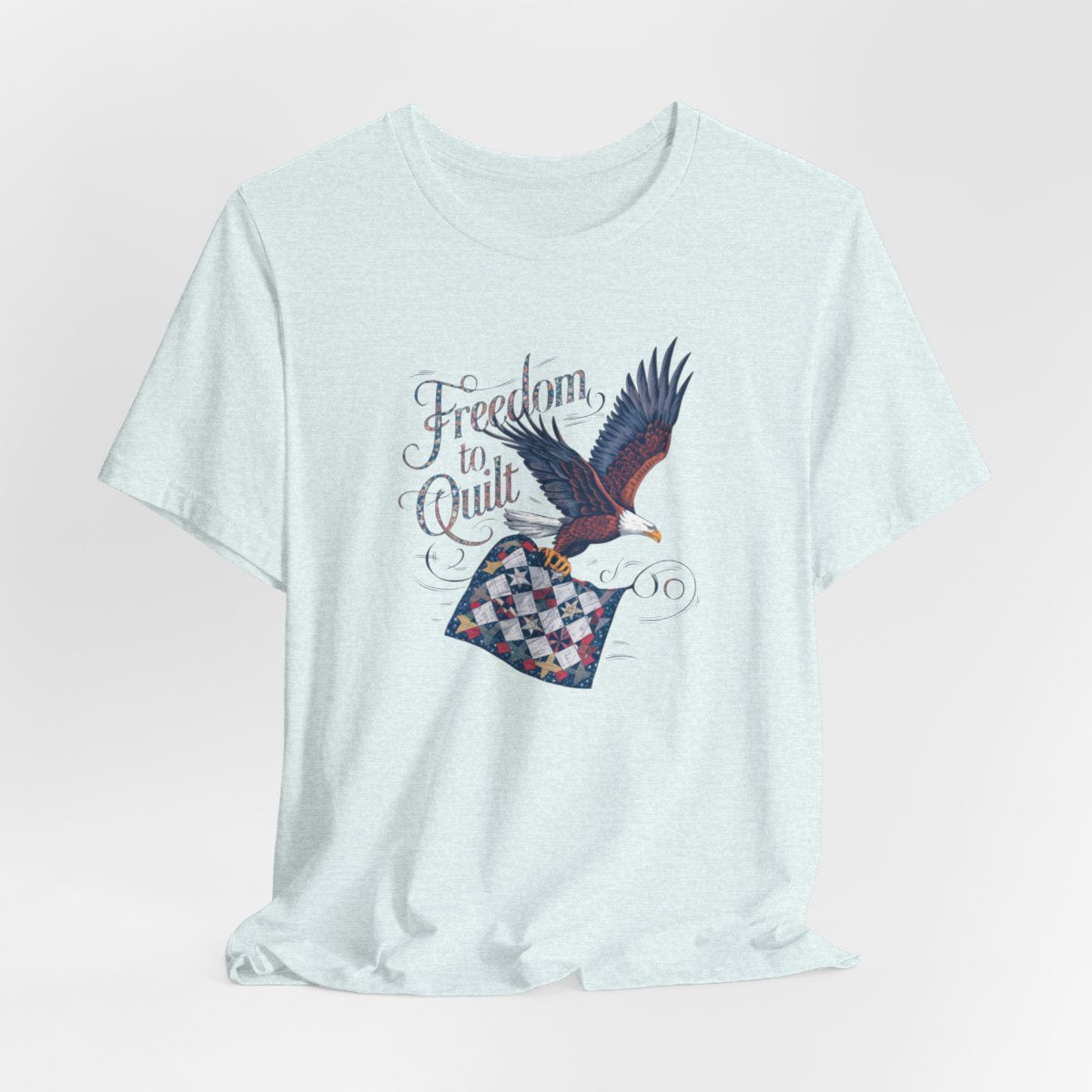 A Heather Ice Blue T-shirt with detailed illustration of a bald eagle in flight, carrying a patchwork quilt. Text reads "Freedom to Quilt" in an ornate, patriotic font.