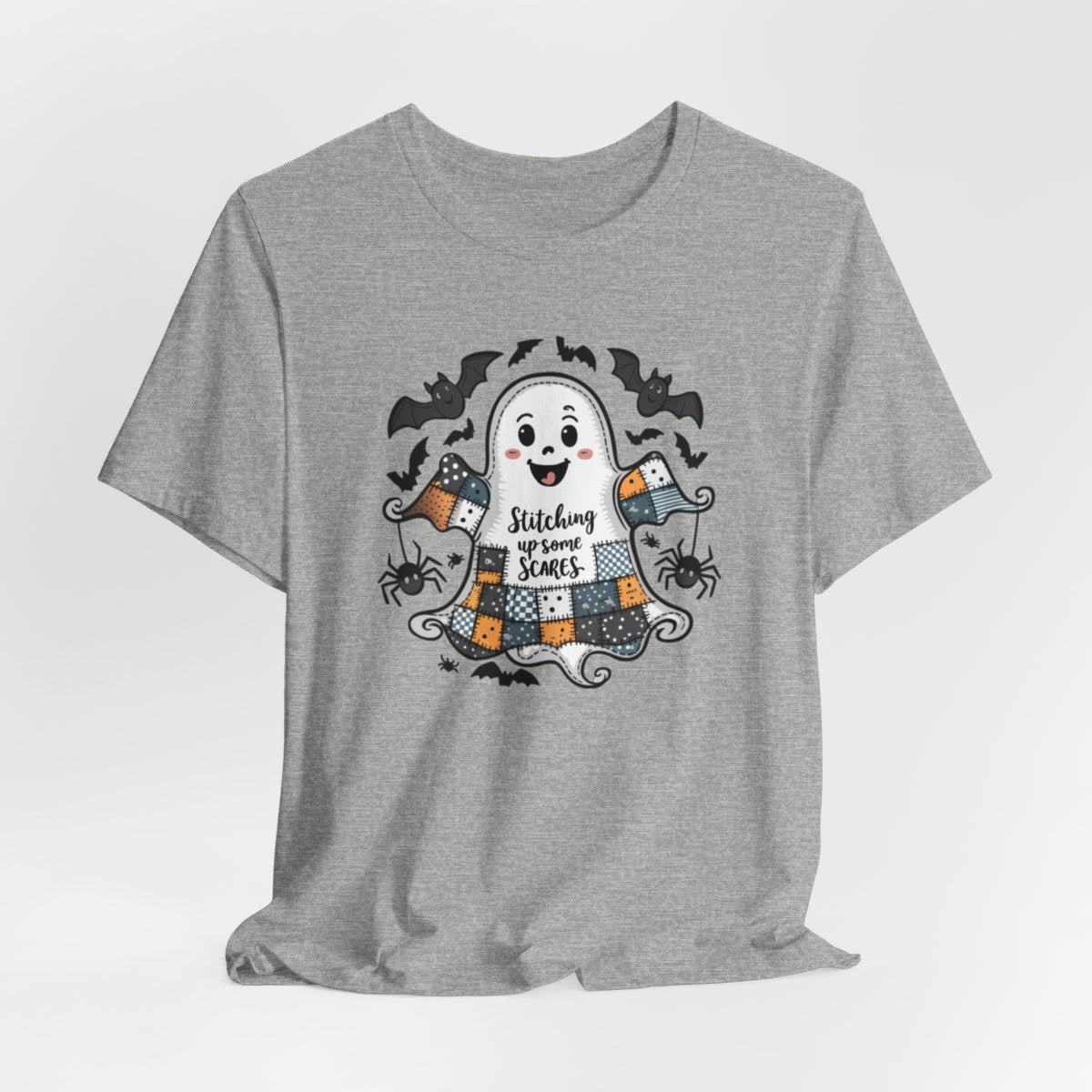 An Athletic Heather Halloween Quilting T-Shirt featuring a smiling ghost with quilted patchwork body. Halloween-themed quilt patterns. Cute bats and spiders around ghost. Text reads "Stitching up some Scares". Ideal for quilters who enjoy Halloween themes.