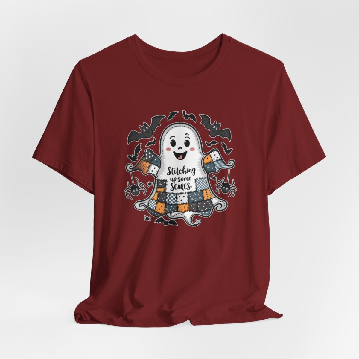 A Cardinal Halloween Quilting T-Shirt featuring a smiling ghost with quilted patchwork body. Halloween-themed quilt patterns. Cute bats and spiders around ghost. Text reads "Stitching up some Scares". Ideal for quilters who enjoy Halloween themes.