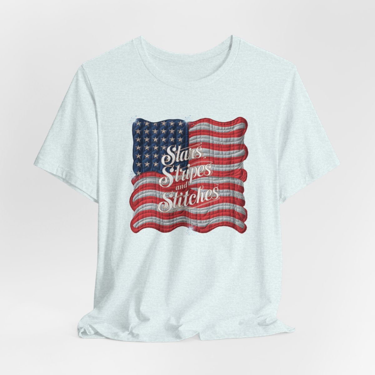 A Heather Ice Blue T-shirt featuring quilted American flag design with text "Stars Stripes and Stitches" overlaid in stylized, patriotic font. Fireworks accent the corners.