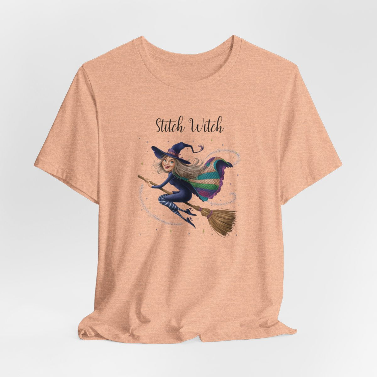 A Heather Peach Halloween Quilting T-Shirt featuring a witch riding broomstick on t-shirt design. Colorful patchwork quilt cape with various patterns. Sparkling magic and stars. Text reads "Stitch Witch". Ideal for quilters who enjoy magical themes.