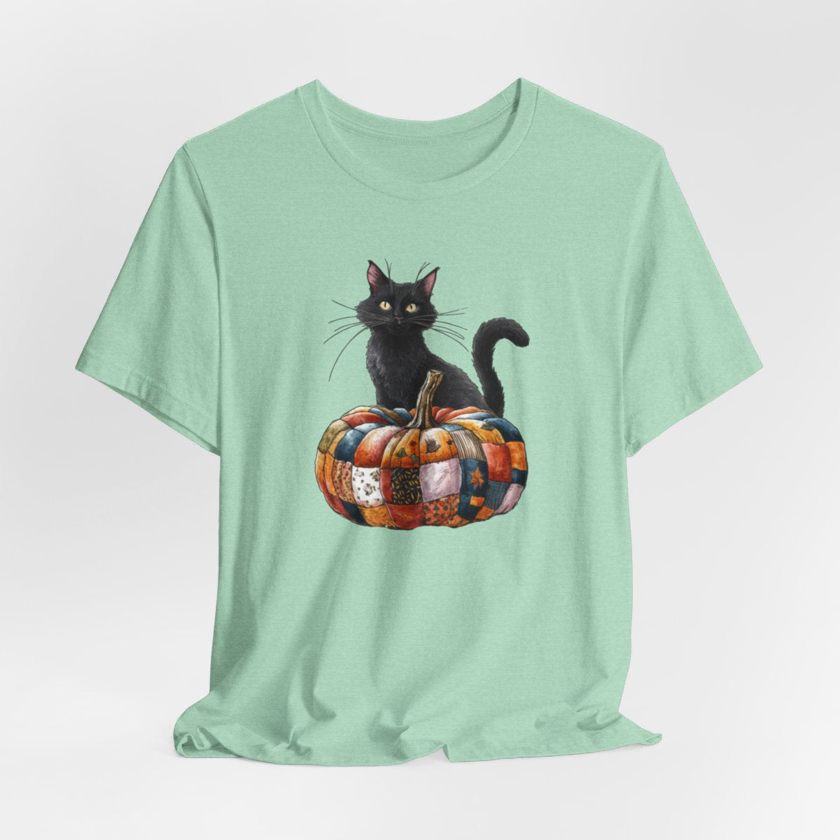 A Heather Mint Halloween Quilting T-Shirt featuring a black cat sitting on colorful patchwork quilt pumpkin. Cat has yellow eyes and long whiskers. Pumpkin features various quilt patterns in autumn colors. Halloween-themed t-shirt design for quilting enthusiasts.